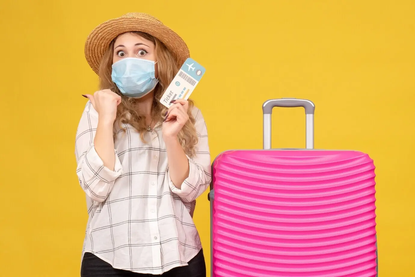 Vorderansicht eines überraschten reisenden Mädchens mit Maske, das ein Ticket zeigt und neben seiner pinkfarbenen Tasche vor gelbem Hintergrund steht.