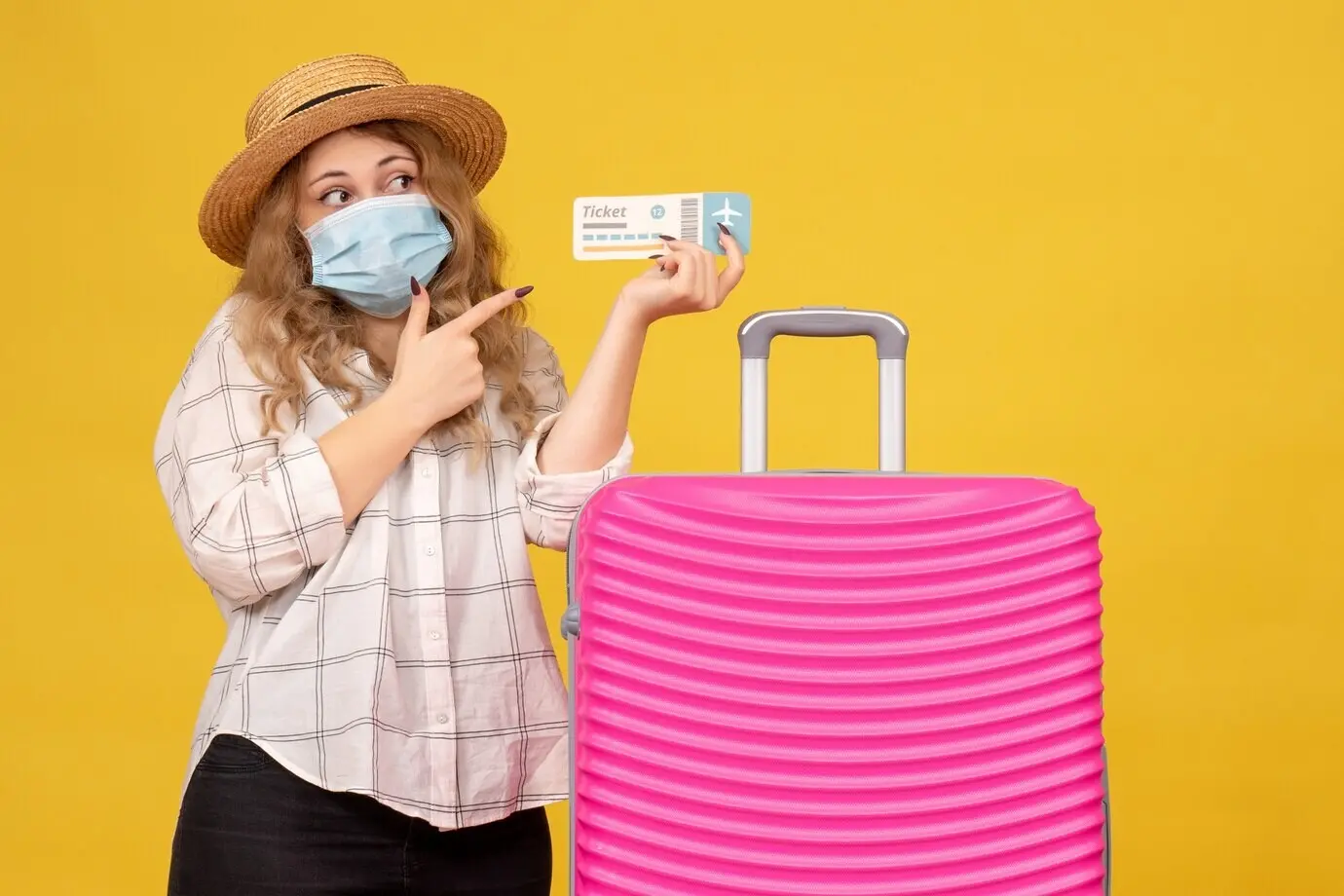 Draufsicht auf eine emotionale junge Frau, die eine Maske trägt, auf ein Ticket zeigt und neben ihrer pinken Tasche steht.